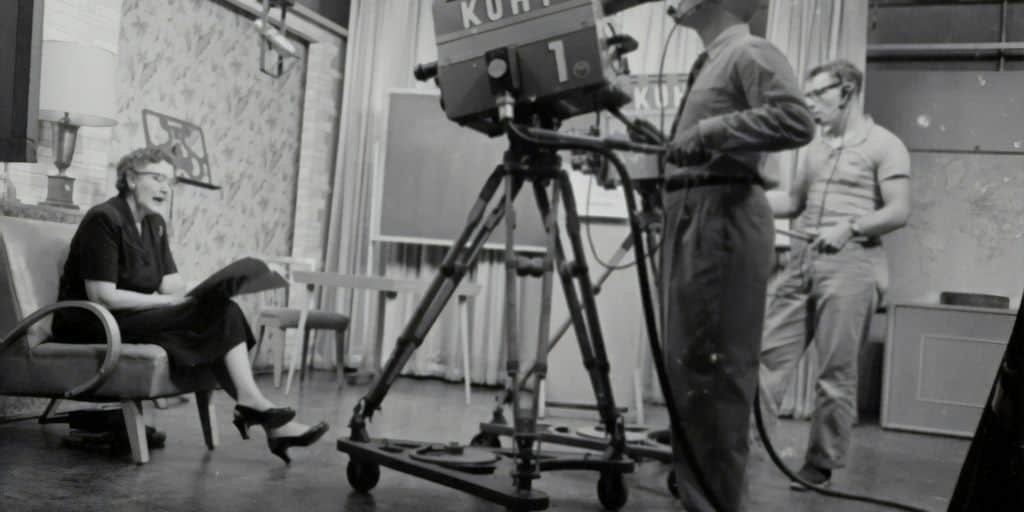 grayscale photography of man standing near studio camera and woman sitting while holding book
